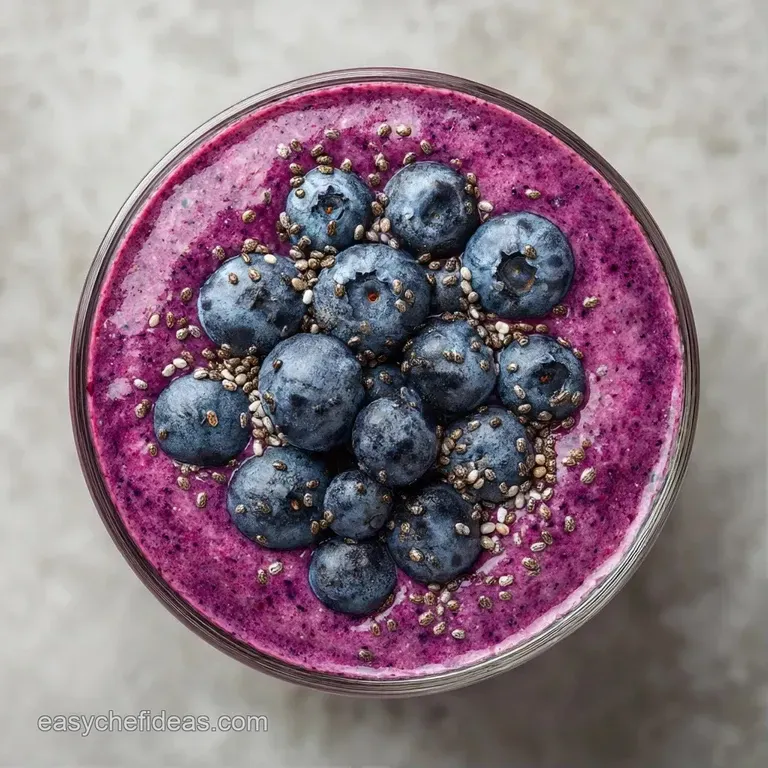 Thick pink smoothie in a tall glass on a wooden tray with a side of fresh blueberries and a mint sprig garnish.